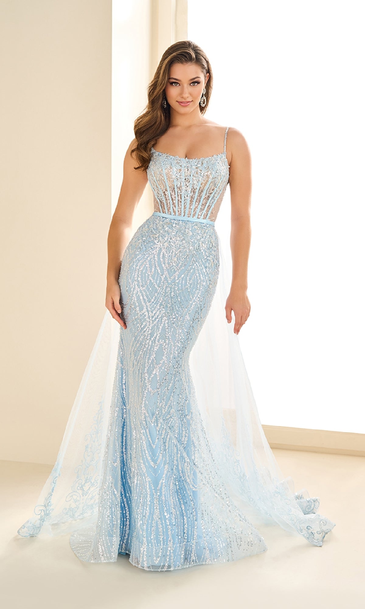 A woman wears the Ellie Wilde EW36075, a fitted light blue sequin prom dress featuring sheer panels, thin straps, and a flowing tulle overlay.